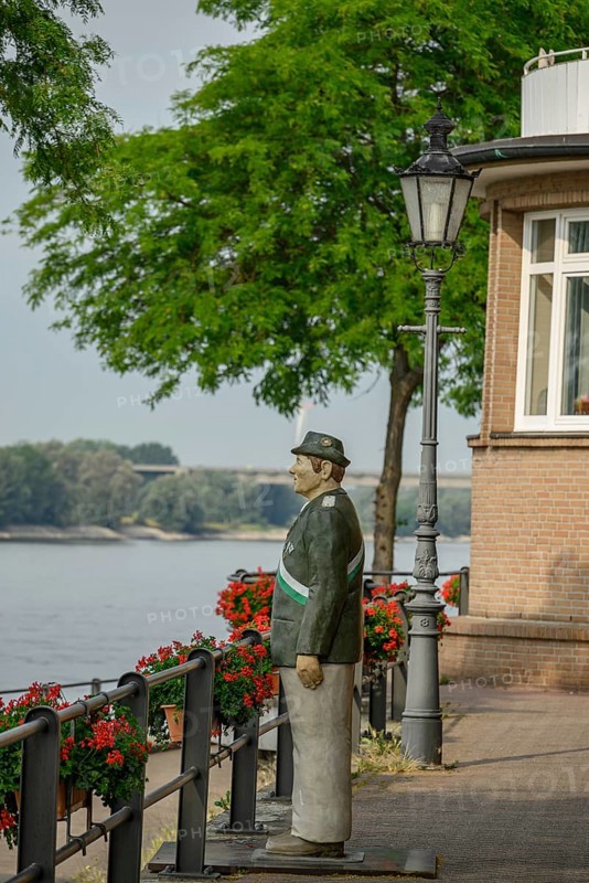 Statue of a man in uniform next to a walkway on the riverbank with lamps and blooming flowers, rhine promenade directly on the river with old houses, lanterns and green trees, rees, germany