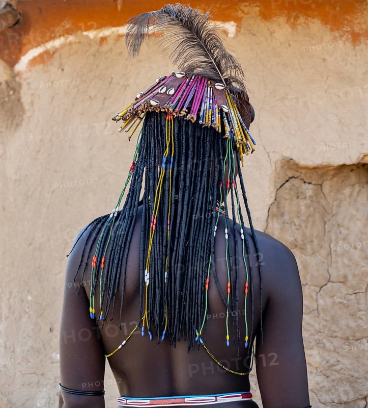 Hakaona woman with traditional kapapo hairstyle and hair ornaments with ostrich feathers, and colourful necklaces, from behind in the morning light, Angolan tribe of the Hakaona, near Opuwo, Kunene, Namibia, Africa