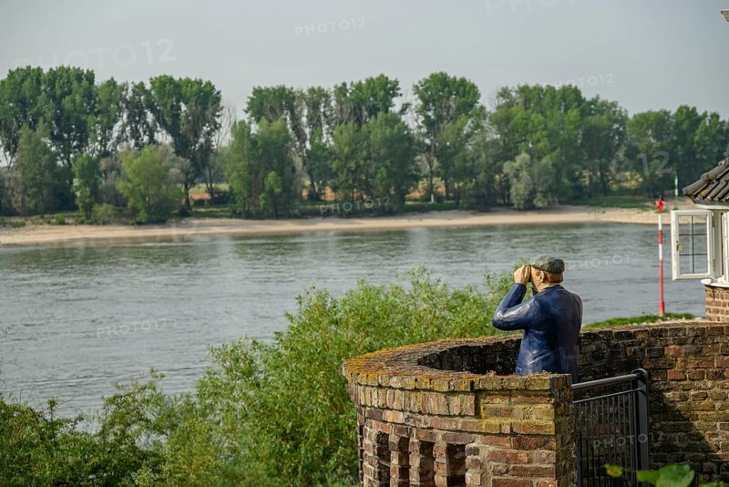 A statue of a sailor with binoculars looking at the riverbank and the trees, rhine promenade directly on the river with old houses, lanterns and green trees, rees, germany