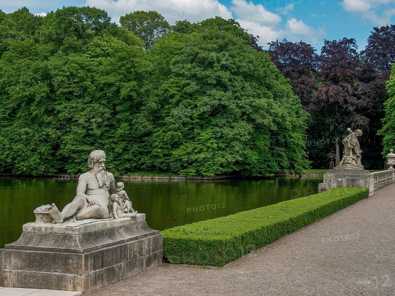 Several stone sculptures along a pond, surrounded by green trees and manicured hedges, old red brick castle with towers and a large park with trees, plants and statues, Nordkirchen, Germany, Europe