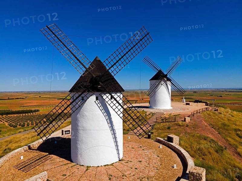 Two windmills stand on a sunny hill in a wide field landscape under a clear sky, aerial view, Alcazar de San Juan, Ciudad Real, Castilla-La Mancha, Spain, Europe