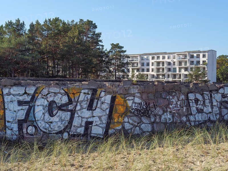 A stone wall with colourful graffiti in front of a modern building, framed by pine trees and tufts of grass, Ruegen, Germany, Europe