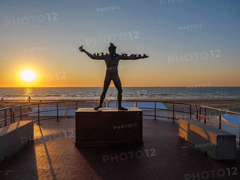 A statue in front of a sunset on the beach, surrounded by a railing, sunset in many colours on the beach with a calm sea, de haan, belgium