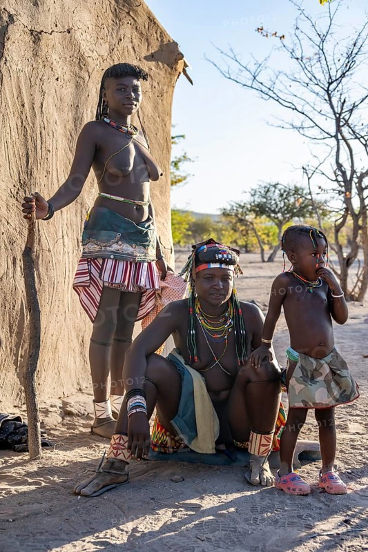 Two Hakaona woman with traditional kapapo hairstyle, with child, in front of their mud hut, in the morning light, in the morning light, Angolan tribe of the Hakaona, near Opuwo, Kunene, Namibia, Africa