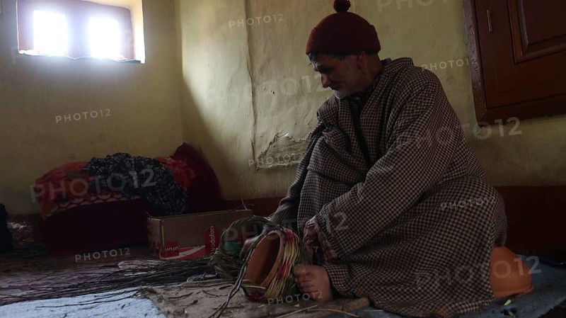 Elderly man in traditional clothing sitting in a dimly lit room with a basket and a contemplative expression, Jammu and Kashmir, India, Asia