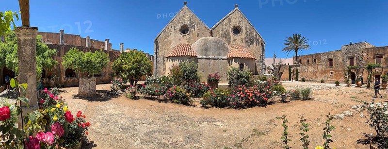 Panoramic photo of monastery courtyard with view of back wall of double-nave church building Church of Arkadi Monastery with double apse in the foreground blooming flowers and shrubs in UNESCO World Heritage Site Orthodox Monastery of Moni Arkadi Arkadi Monastery from early Middle Ages, important national monument national shrine on island of Crete, Arkadi, Armnatos, Crete, Greece, Europe