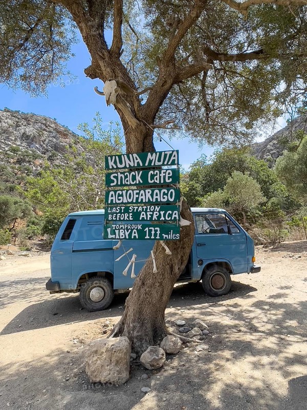 Tourist humorous funny sign on tree behind old Volkswagen VW Bus Bulli in front of entrance of Agiofarago Gorge of Saints on south coast of island Crete with text Last Station in front of Africa, Libya 177 miles, Agiofarago Gorge, Crete, Greece, Europe