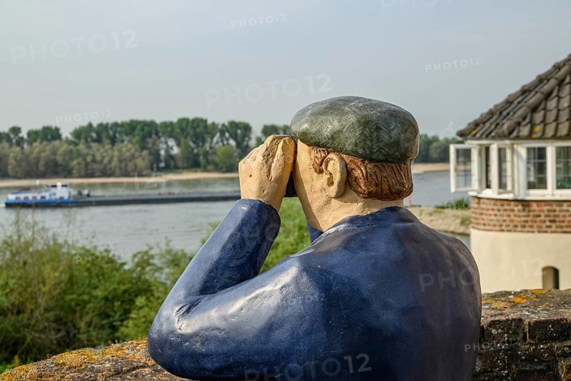A statue of a sailor with binoculars looking at a river and a boat, next to a building, rhine promenade directly on the river with old houses, lanterns and green trees, rees, germany