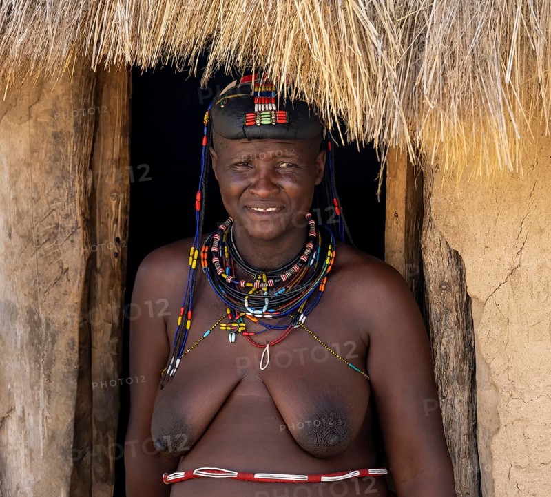 Hakaona woman with traditional kapapo hairstyle, in the entrance of her mud hut, portrait, in the morning light, Angolan tribe of the Hakaona, near Opuwo, Kunene, Namibia, Africa