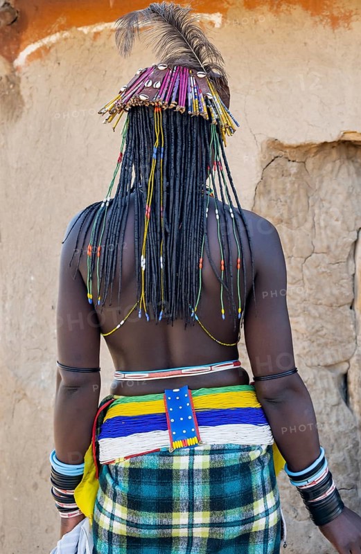 Hakaona woman with traditional kapapo hairstyle and hair ornaments with ostrich feathers, and colourful necklaces, from behind, in the morning light, Angolan tribe of the Hakaona, near Opuwo, Kunene, Namibia, Africa
