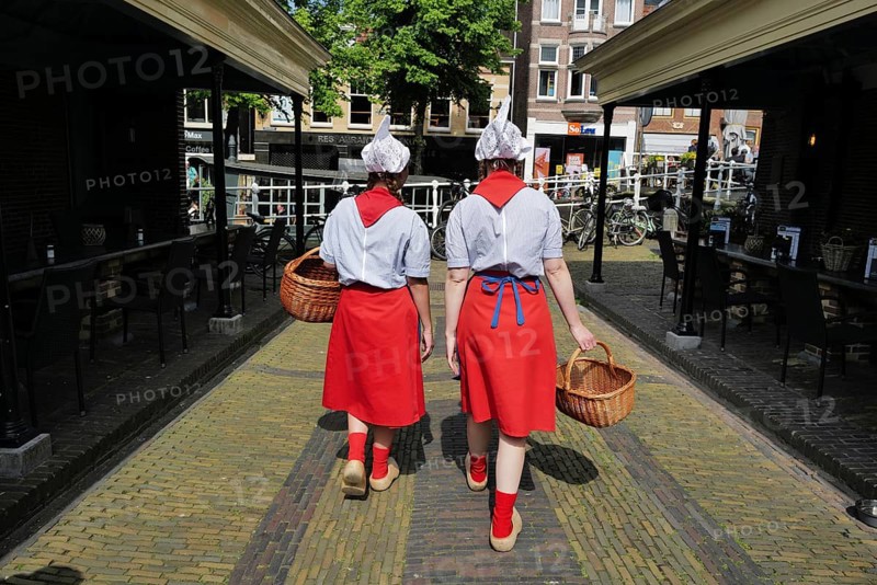 Two Dutch cheese girls in traditional tracht and carrying baskets of cheese, wander through the town of Alkmaar on market day., Alkmaar, The Netherlands, Europe
