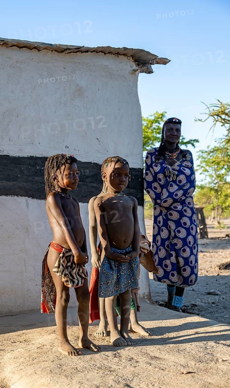 Hakaona woman and two traditional Hakaona children in front of their mud hut, in the morning light, Angolan tribe of the Hakaona, near Opuwo, Kunene, Namibia, Africa