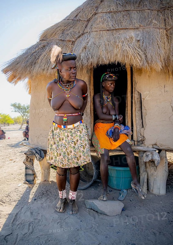 Two Hakaona woman with traditional kapapo hairstyle and hair ornaments with ostrich feathers, in front of their mud hut, in the morning light, Angolan tribe of the Hakaona, near Opuwo, Kunene, Namibia, Africa