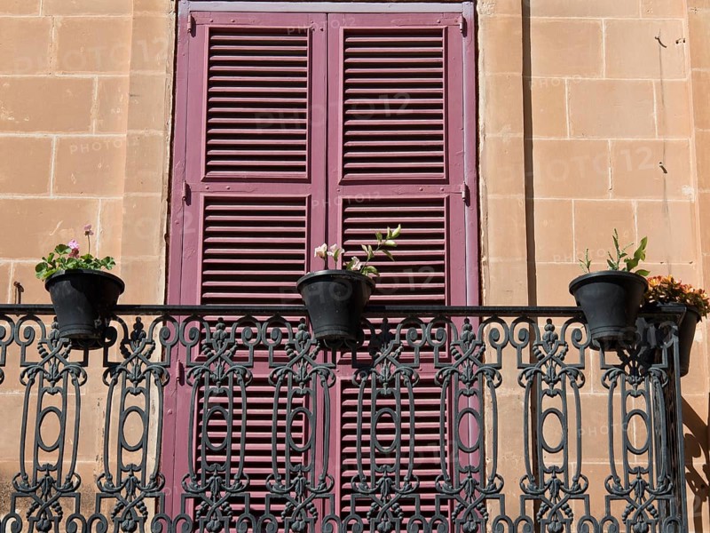 A balcony with wrought-iron railings and pink shutters, decorated with several flower pots in front of a sandstone façade, mdina, mediterranean sea, malta