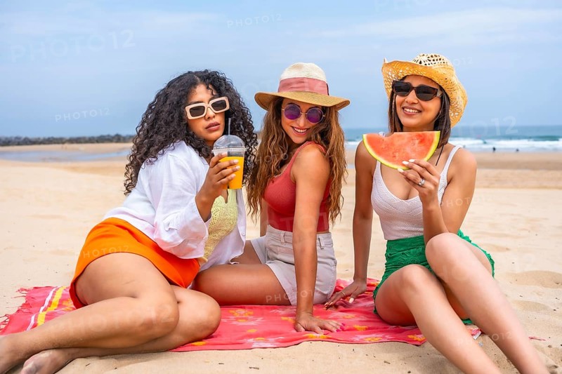 Three women sitting on a beach, one of them holding a watermelon slice. Scene is relaxed and carefree, as the women are enjoying their time together on the beach