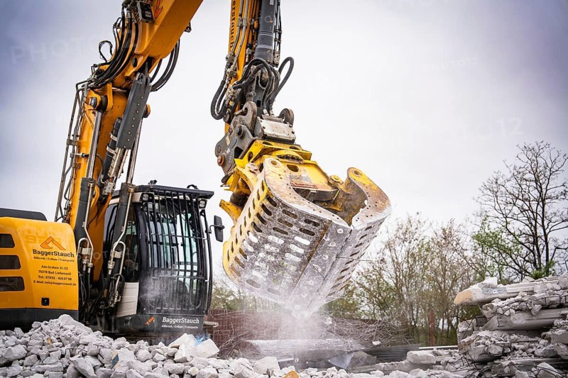 Large excavator with a metal grab crushes building rubble while water splashes and stirs up dust, demolition site, Saarbrücken, Germany, Europe