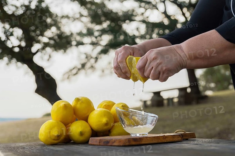 Woman squeezes a lemon with her hands the juice falls into a glass bowl, in the background is a pile of lemons and forest trees