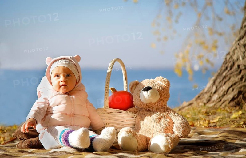 Baby sitting on a blanket outdoors with a teddy bear and basket containing a pumpkin on a sunny fall day by the lake, Belarus.Minsk