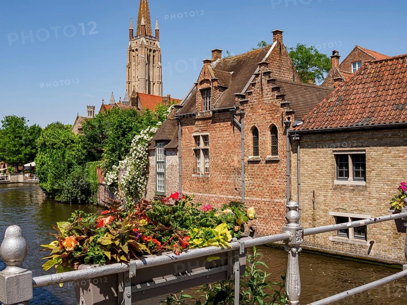 A canal with decorated balustrades and flowers along the row of brick-built houses in front of a church tower in sunny weather, Bruges, Belgium, Europe