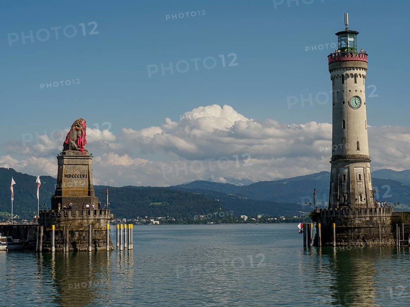 Lion statue and lighthouse with clear sky and calm water, nearby town and mountains in the background, Lindau, Bavaria, Germany, Europe