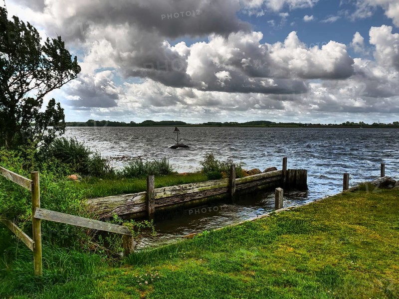 Lake view with jetty and lush shore grass under a dramatic sky with dense clouds, Maasholm, Schleswig-Holstein, Germany, Europe