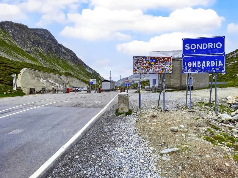 View from Switzerland towards Italy of the former border crossing between Switzerland and Italy at the top of the Splügen Pass Passo dello Spluga on the right blue sign with white lettering for the province of Sondrio below sign for the region of Lombardy Lombardia next to it brown sign Splügen Pass, Canton of Graubünden, Switzerland, Lombardy, Italy, covered with stickers by tourists, Europe