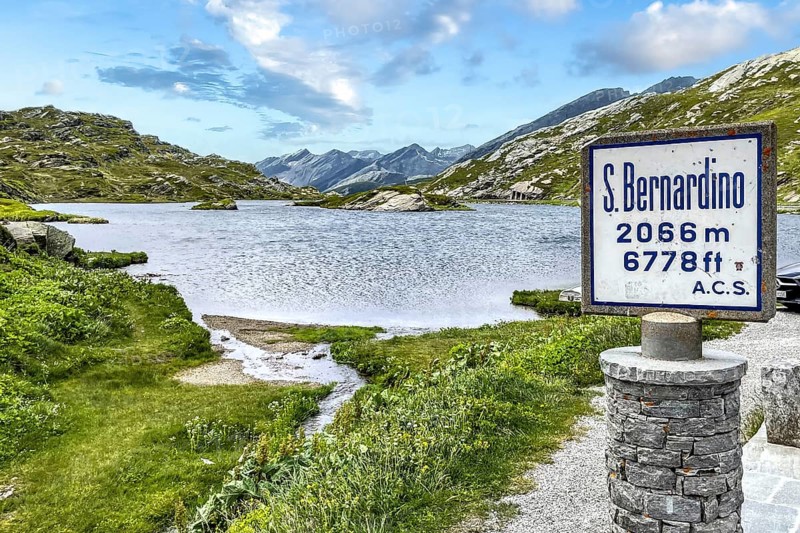 Pass marker sign at pass summit of mountain pass in Alps Passo San Bernardino San Bernardino Pass 2066 metres m 6778 feet ft high Alpine pass Alpine road Pass road, behind it small mountain lake Lake Laghetto Moesola, Canton Graubünden, Switzerland, Europe