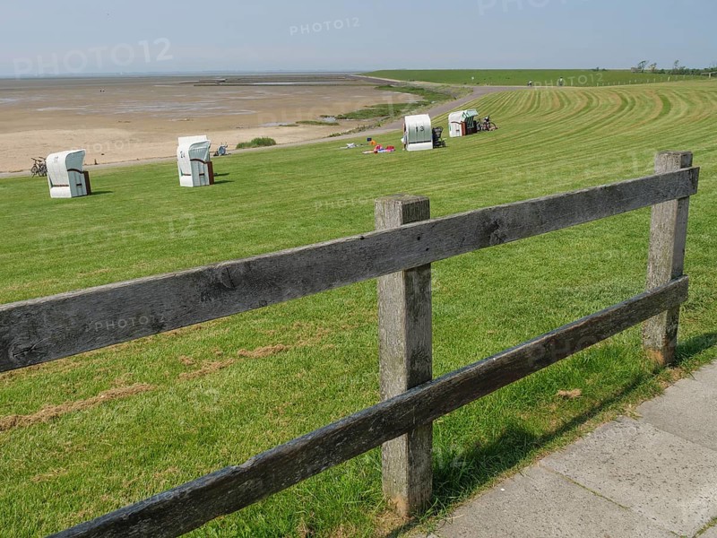 Meadow with beach chairs and a wooden fence along a path in front of a vast sandy beach and the sea under a clear sky, hallig hooge, schleswig-holstein, germany