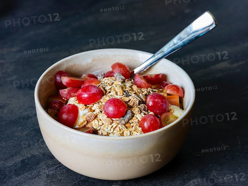 Fresh homemade muesli with organic red grapes and oat flakes in a ceramic bowl with a spoon on a dark-coloured base