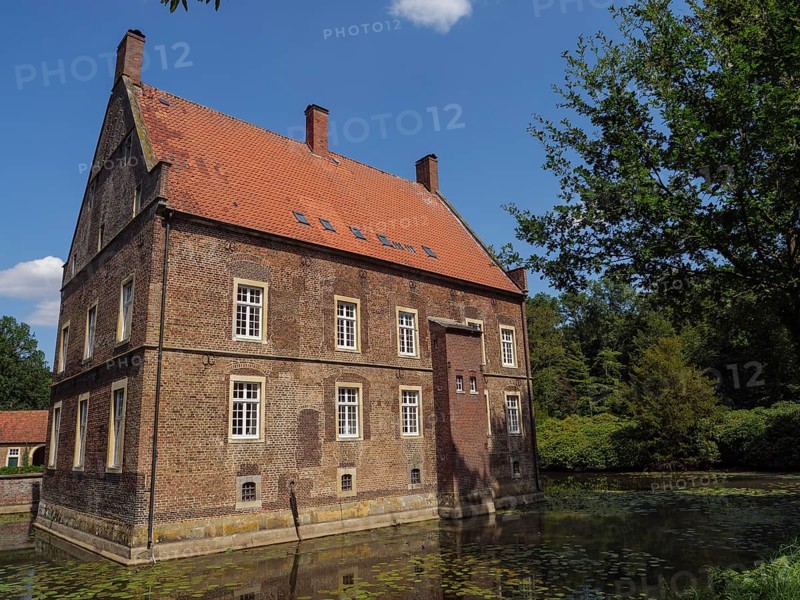 Old brick house with red roof on the edge of a pond, surrounded by green trees in summer, ochtrup, münsterland, germany