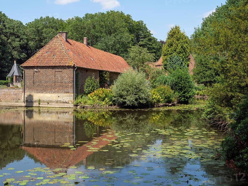 Brick building by a pond with a clear reflection, surrounded by trees and green vegetation on a sunny day, ochtrup, münsterland, germany