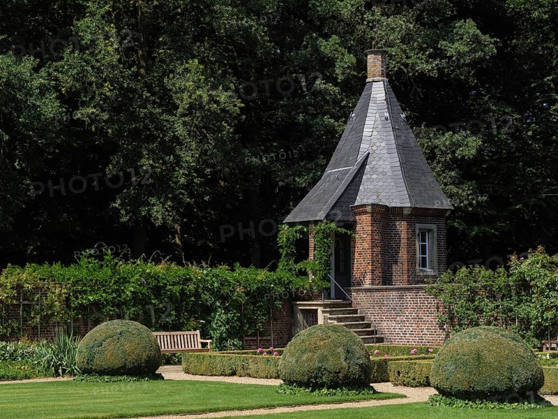 A small tower with a slate-covered roof in the middle of a well-tended garden with shrubs, green grass and trees providing shade, ochtrup, münsterland, germany