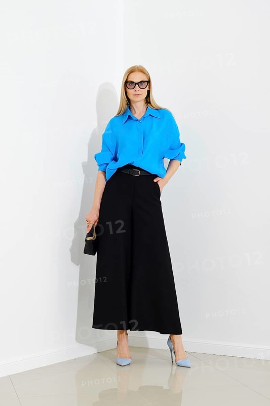 Confident woman in blue button down shirt and black maxi skirt posing over white wall