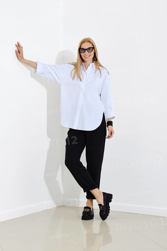 Woman in white cotton shirt and black trousers leans her hand against the wall while standing in a bright room with white walls