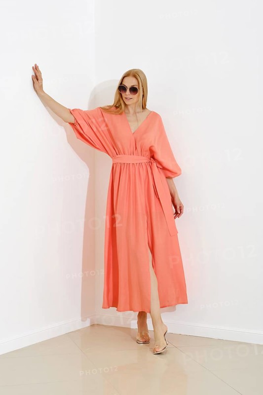 A stylish blonde woman in a coral maxi dress with a v-neckline leaning against white wall