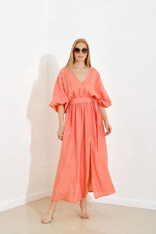 A full-length portrait of a woman in a coral maxi dress with a v-neckline and short sleeves, cinched at the waist, standing against white wall