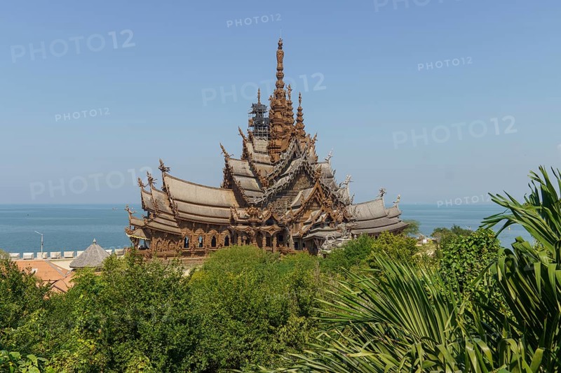 Wooden temple with intricate architecture surrounded by lush greenery, with the sea and blue sky in the background, Pattaya, Thailand, Asia