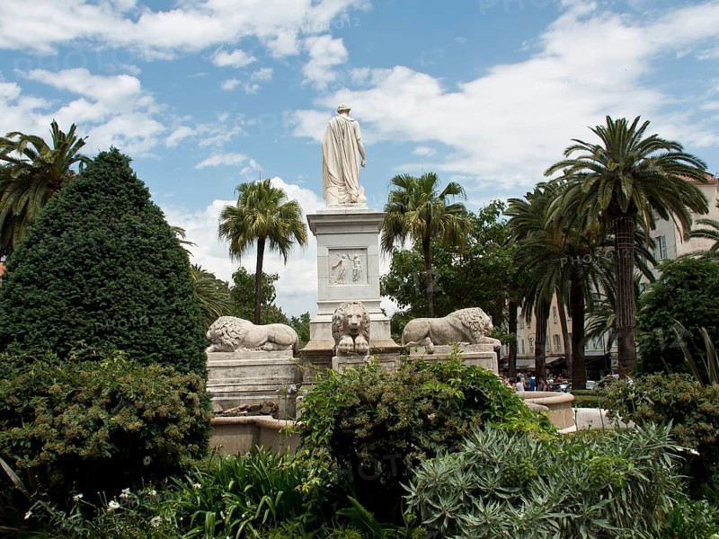 An impressive statue of a lion in a fountain surrounded by palm trees in a park under a cloudy sky, ajaccio, corsica, france