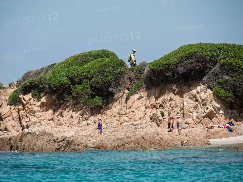 People walking along a rocky coastline, overgrown cliffs in the background and blue sea, ajaccio, corsica, france