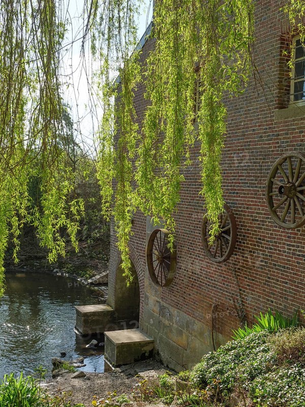 A brick building with large wooden wheels sits on the riverbank, surrounded by green trees and overhanging branches on a sunny spring day, metelen, westphalia, germany