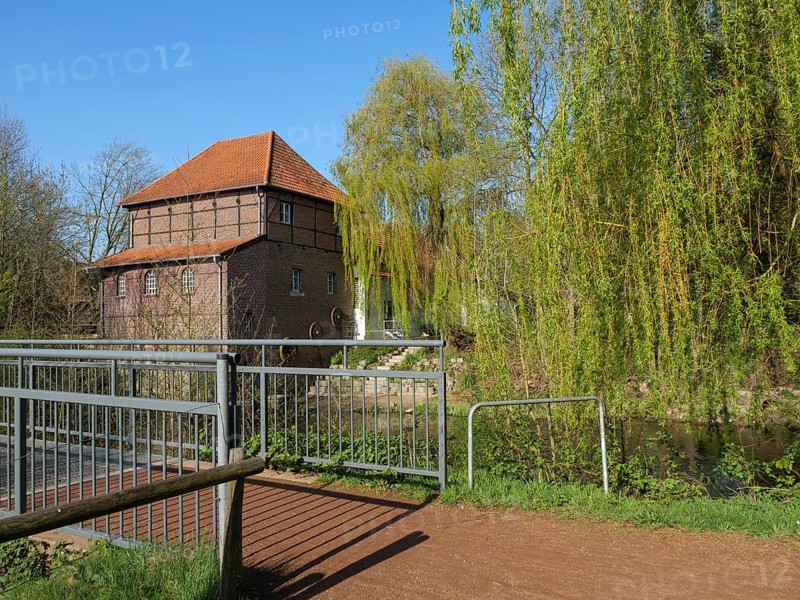 A narrow path with a bridge and metal railings leading to a brick building with a red roof, surrounded by green nature and a clear sky, metelen, westphalia, germany