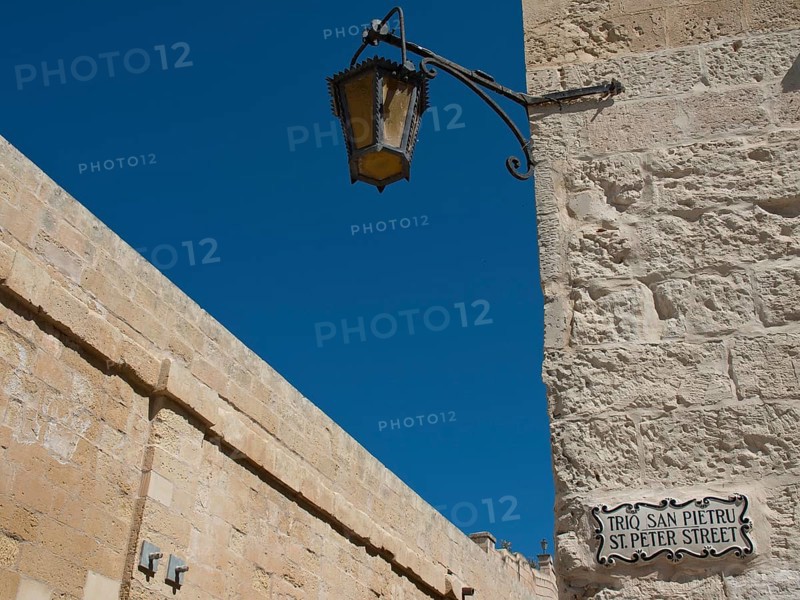 A decorative lamppost and street sign along an old stone wall under a blue sky, mdina, mediterranean sea, Malta, Europe