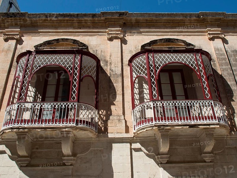 Two ornate and decorative iron balconies in front of a historic stone wall with windows, mdina, mediterranean sea, malta