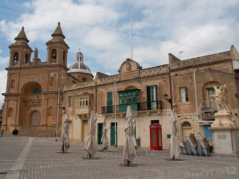 A historic square with a large church and surrounding buildings under a cloudy sky, marsaxlokk, mediterranean sea, malta