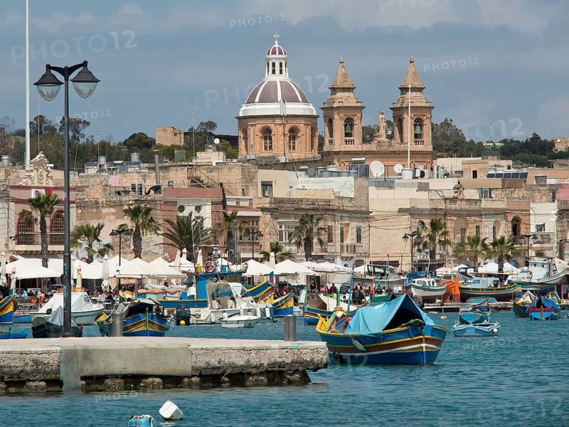 Fishing boats in the harbour with a cathedral in the background under a cloudy sky, marsaxlokk, mediterranean sea, malta