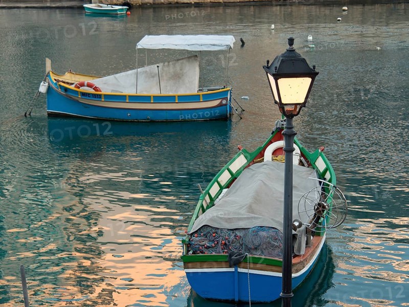 Two colourful fishing boats floating on a calm waterway with a lantern in the foreground, marsaxlokk, mediterranean sea, malta