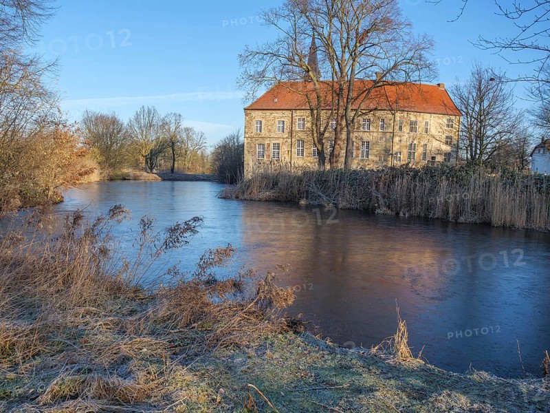 Historic building with red tiled roof on the riverbank, surrounded by bare trees in a wintry ambience, lüdinghausen, westphalia, germany