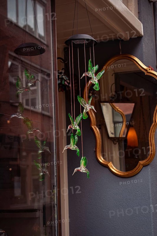 Glass birds in shades of green hang from a ceiling and are reflected in a window. A golden, antique mirror is attached to the wall behind it, Amsterdam, Netherlands