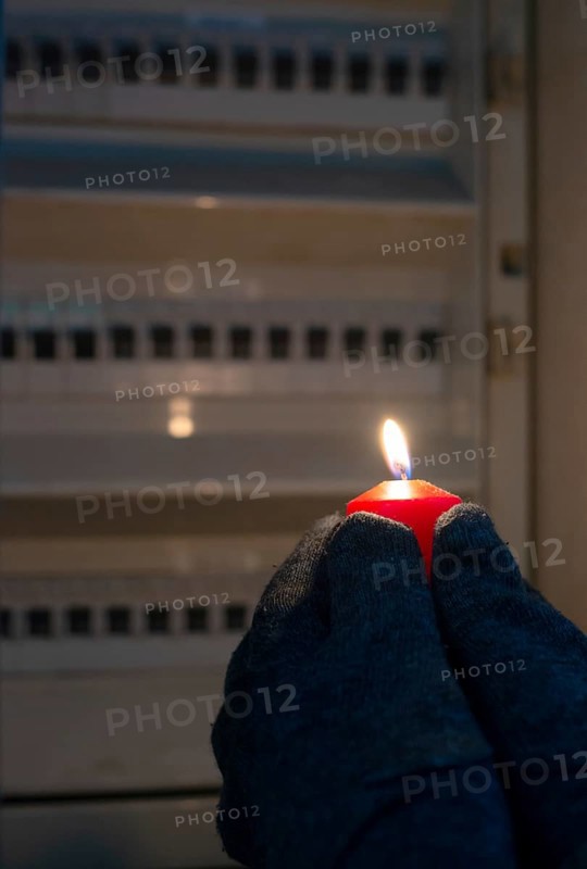 Blackout, standing with a candle in front of a power fuse box, energy cut out, uncertain supply, risk and crisis for electricity