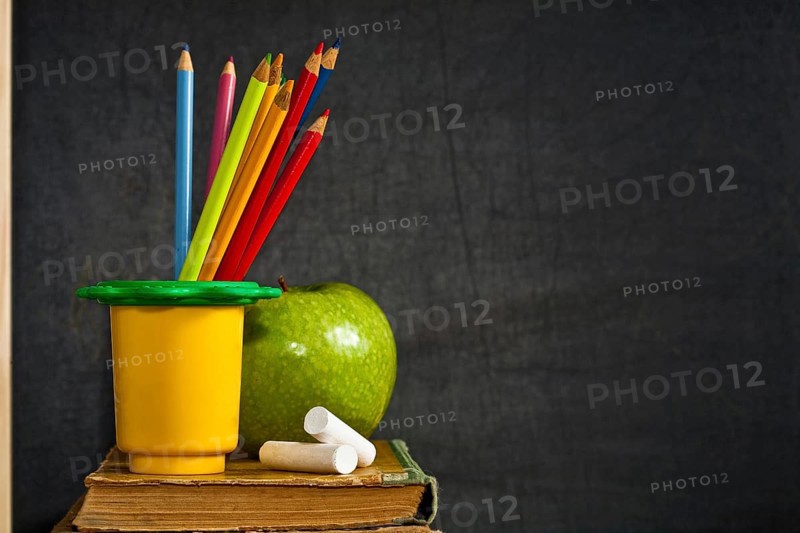 Multicolor pencil, chalk and green apple on old textbook against blackboard in class. School concept